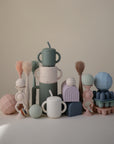 A pastel arrangement of mushie Snack Cups with handles and lids is displayed among abstract geometric shapes—spheres, arches, and brush-like forms—set against a neutral background.