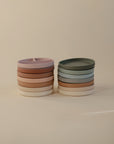 Two stacks of mushie Silicone Suction Plates in pastel pink, tan, green, blue, and white are arranged against a beige background. One plate features two compartments ideal for baby feeding.