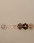 Six FRIGG Natural Rubber pacifiers in neutral shades, from light beige to dark brown, are arranged in a row against a plain light beige background. Made from natural rubber latex in Denmark.