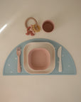 The mushie Dinnerware Cutlery Set features a pink plastic fork, knife, bowl, and cup on a pastel blue semicircular placemat with a beige teether toy placed above the utensils—all arranged neatly on a light surface.