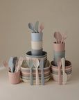 The mushie Dinnerware Cutlery Set features pastel bowls, plates, and cups, each with non-toxic plastic cutlery in pink, blue, green, and cream tones, all arranged neatly on a neutral background.