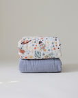 A mushie Organic Cotton Muslin Swaddle Blanket with a space-themed planets and stars print is stacked on a plain light blue baby blanket, both neatly folded against a simple, light background.