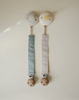 Mushie Linen Pacifier Clip 2-Pack: blue & beige pacifiers with wooden clasps on light background.