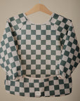 A mushie Long Sleeve Bib featuring a green and white checkered pattern and water-resistant fabric hangs on a wooden hanger against a plain beige background.