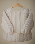 The mushie Long Sleeve Bib features water resistance, elastic cuffs, light pastel vertical stripes, and a wide bottom pocket. Displayed on a wooden hanger against a neutral backdrop.