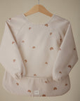 The mushie Long Sleeve Bib, water-resistant and light beige with elastic cuffs and brown rainbow patterns, hangs on a wooden hanger against a plain background.