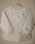 The mushie Long Sleeve Bib in light beige features water-resistant fabric, elastic cuffs, and a small bee pattern. It hangs on a wooden hanger against a plain background.