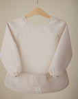 The mushie Long Sleeve Bib in light beige, featuring elastic cuffs and water-resistant, wipe-clean fabric, is displayed on a wooden hanger against a neutral background. A small tag accents the bottom center of its simple design.