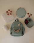 Flat lay of a light green backpack with a wooden beaded toy, baby bottle, folded white cloth, silicone suction cups, and the mushie Lunchbox—pale green, non-toxic plastic with built-in compartments—plus a divided snack container filled with cherries.