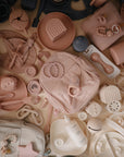 A flat lay of assorted baby essentials in soft pink, beige, and neutrals, including the mushie Silicone Baby Rattle Toy, pacifiers, teethers, bottles, plates, utensils, bibs, and toys—all neatly arranged on a light background.