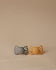 The FRIGG Lucky Symmetrical Silicone Pacifier 2-Pack (6-18 Months) by FRIGG features gray and mustard yellow pacifiers with butterfly-shaped handles, displayed side by side on a smooth beige surface with a matching background.