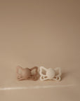 The FRIGG Butterfly Anatomical Silicone Pacifier 2-Pack (6-18 Months) by FRIGG is shown side by side on a beige background, featuring one light brown and one white pacifier with butterfly-shaped handles and round nipples.