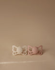 The FRIGG Butterfly Anatomical Silicone Pacifier 2-Pack (6-18 Months) by FRIGG features one white and one pink butterfly-shaped pacifier, shown side by side on a light beige surface with a matching background.