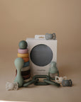 The mushie Silicone Baby Rattle Toy, made from food-grade silicone, is shown alongside stacking rings, pacifiers, a teether, and a boxed set—all displayed on a beige surface with a soft beige background for safe sensory play.