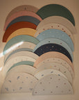 Sixteen mushie Silicone Placemats in assorted pastel colors and patterns are neatly overlapped on a beige wall, with sunlight casting a bright diagonal streak across the display.
