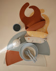 A flat lay of mushie BPA-free silicone baby feeding items, including two bibs (brown and patterned yellow), the Round Dinnerware Bowl, plates, a cup, utensils, and semi-circle placemats in muted colors on a cream background.
