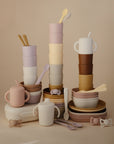 A stylish display of mushie Dinnerware Cups in pastel beige, pink, brown, and cream towers above matching bowls, plates, and spoons. These non-toxic kids’ cups and dinnerware sit against a soft beige background.