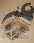 A flat lay featuring two silicone bibs, a mushie Silicone Placemat, suction bowl, plate, cup, and toddler utensils in muted blue and beige with playful animal and star patterns—all crafted from food-grade silicone.