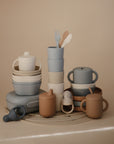 Mushie Dinnerware Cups and other pastel silicone baby feeding items—non-toxic cups, bowls, plates, fork, spoon, and pacifier—are neatly arranged on a beige surface as perfect alternatives to plastic dinnerware.