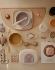 A neatly arranged collection of pastel mushie baby feeding items and toys, featuring bowls, cups, spoons, a teether, a bib, food-grade silicone accessories, and the mushie Silicone Baby Rattle Toy for added sensory experience on a beige background.