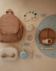 A flat lay of pastel baby items from mushie, featuring a tan backpack, a blue and tan silicone plate, utensils, tea set, pacifier, paint palette toy, a white container, and the Silicone Baby Rattle Toy—neatly arranged on a beige background.