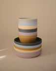 A stack of mushie Round Dinnerware Bowls and plates in yellow, pink, gray, and light blue—BPA-free pastel-colored plastic—arranged neatly against a plain beige background.
