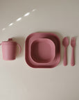 The mushie Square Dinnerware Plates set in pink includes BPA-free plastic plates, a sippy cup, spoon, and fork, all neatly arranged on a light beige surface.