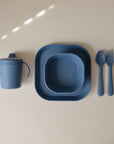 The mushie Dinnerware Cutlery Set features matte blue, non-toxic silicone children's tableware—a sippy cup with handles, bowl, plate, fork, and spoon. Dishwasher-safe and neatly arranged on a beige surface.