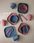 Four mushie Dinnerware Fork and Spoon Sets in blue and pink are displayed on a beige surface. Each set includes dishwasher-safe silicone plates, handled cups, forks, spoons, matching utensils, and a sippy cup.