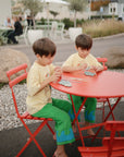 Two kids outdoors play with mushie’s Silicone Sensory Toy at a red table for fine motor fun.