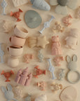 A flat lay of pastel mushie Daisy Silicone Suction Builders and other baby toys—stacking cups, castles, bunny teethers, and animal figures—on a neutral beige background to encourage motor skills development.