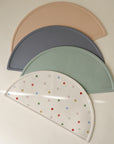 Four semi-circular mushie Silicone Placemats—beige, gray, green, and white with colorful dots—are stacked on a neutral surface under soft light. Made from food-grade silicone, they're safe and vibrant for any meal.