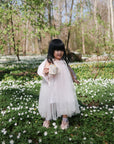 A toddler in a light pink dress stands among white wildflowers, holding a mushie Snack Cup. Green foliage and trees surround her as she enjoys a sunny day.