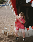 A young girl smiles happily on the playground beside her mushie Mini Toddler Bike.