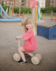 Young girl in pink rides a beige mushie Mini Toddler Bike at the playground, smiling happily.