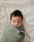 Baby with dark hair wrapped in mushie Organic Cotton Muslin Swaddle Blanket, on white cloth.