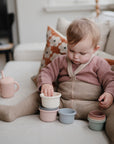 A toddler in a pink sweater, taupe tights, and brown shorts stacks mushie Stacking Cups Toy on the couch. A pink sippy cup sits nearby, with a floral pillow in the background.