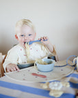 A young blonde child sits in a highchair, eating spaghetti with a fork. The table features a mushie Silicone Placemat and matching utensils, while the child wears a bib and dines on a blue and white striped tablecloth.