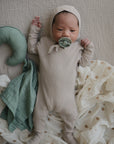 A baby in a beige ribbed onesie and hat lies on a textured blanket, holding a green pacifier. Beside the baby are a green crescent pillow and the mushie Moon Lovey Blanket, crafted from organic cotton for added comfort.