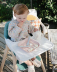 A toddler sits outdoors on a high chair, wearing a floral bib and eating cut fruit from a pink mushie Square Dinnerware Plate, holding a cup with both hands amid greenery and wooden furniture.