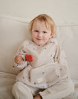 A young child with light hair smiles on a beige couch, holding a red popsicle made with the mushie Toddler Ice Pop Tray. The child wears a long-sleeved, light smock adorned with small sailboat patterns.