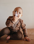 Smiling baby holds mushie Figurine Teether on wooden floor, in floral top and brown pants.