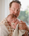 A man with closed eyes gently holds a sleeping newborn with a FRIGG Natural Rubber pacifier from the FRIGG 6-Pack, wrapped in a light blanket against his chest in a softly lit room.