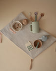 The mushie Silicone Toddler Starter Spoons, in soothing pastel colors, are arranged neatly on a beige ribbed cloth—paired with bowls, cups, a toy phone, and pacifiers—creating an inviting setup for independent feeding.