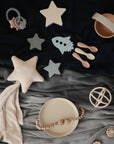 Flat lay of neutral-toned mushie baby items, featuring star-shaped teethers, a rocket teether, silicone spoons, suction bowl and plate, pacifier clip, Star Lovey Blanket, organic cotton muslin cloth, and a ring rattle.