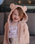 Smiling child in mushie's Bear Robe over a floral swimsuit, sitting on a couch.
