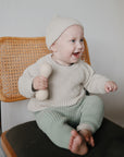 A smiling baby in a beige knit hat and sweater with green pants sits on a wooden chair, holding the mushie Silicone Baby Rattle Toy made from food grade silicone for safe and soothing play.