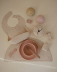 The mushie Snack Cup is shown with pastel baby and toddler feeding items—a speckled bib, divided placemat, bowl and spoon, cup, toy cupcakes, rattle, and pacifier clip—arranged on a light surface.