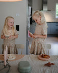 Two young girls with blonde hair in matching floral dresses stand by a dining table set with candles, plates, snacks, and a mushie Silicone Placemat made from food-grade silicone, smiling and interacting in a cozy modern kitchen.