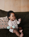 A smiling baby with curly hair sits on a dark green couch, holding a food-grade silicone pacifier and the mushie Silicone Baby Rattle Toy in pale blue, with a patterned cushion in the background.